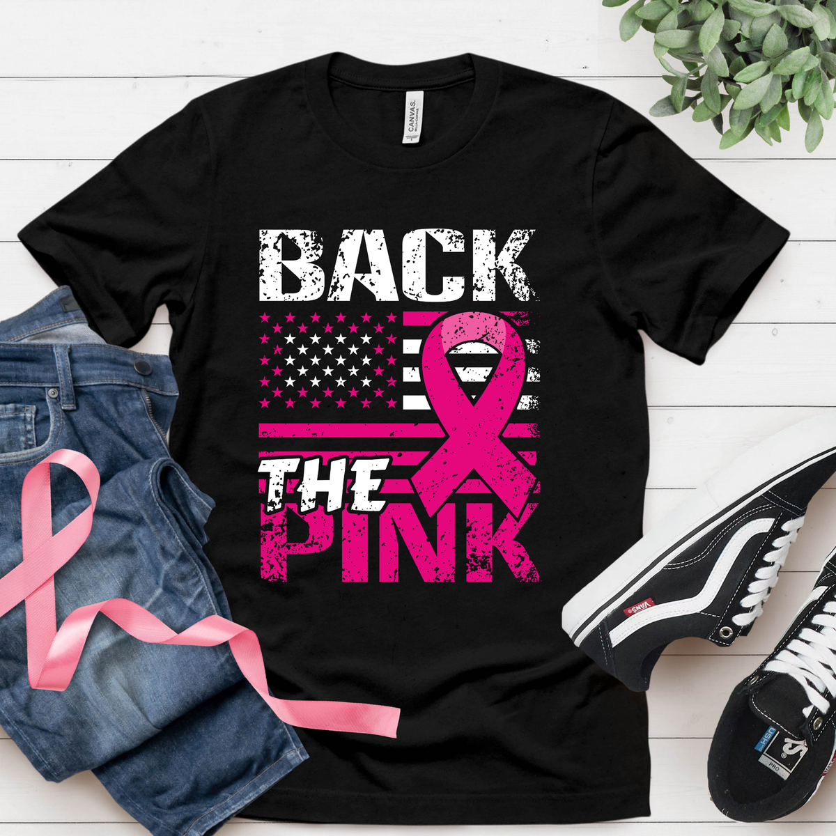 Black t-shirt with pink ribbon design and text on a white wooden surface with jeans and sneakers.