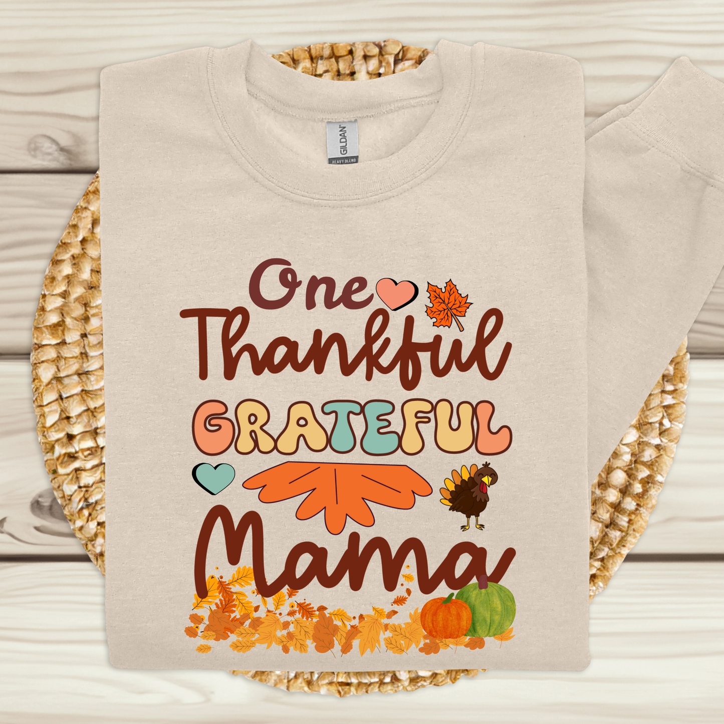 Beige sweatshirt with 'One Thankful Grateful Mama' text and fall-themed graphics on a woven mat.