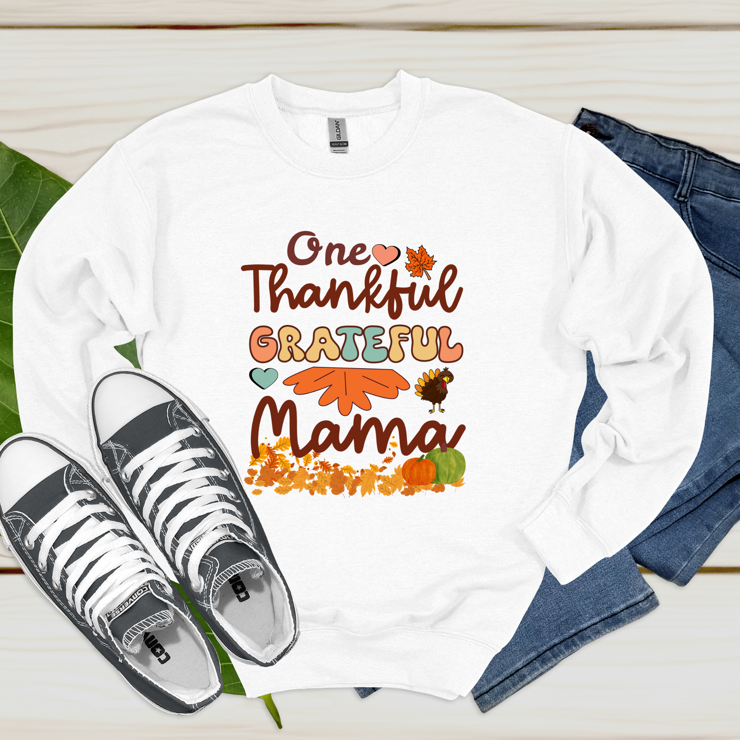 White long-sleeve shirt with 'One Thankful Grateful Mama' text and fall-themed graphics, paired with sneakers and jeans on a wooden surface.