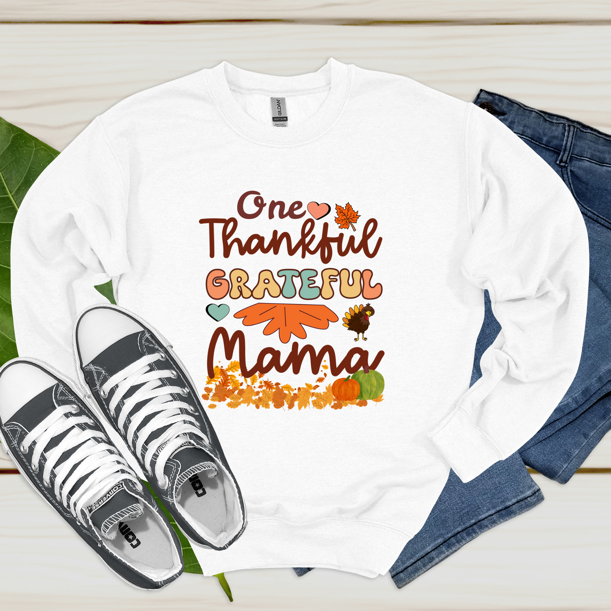 White long-sleeve shirt with 'One Thankful Grateful Mama' text and fall-themed graphics, paired with sneakers and jeans on a wooden surface.