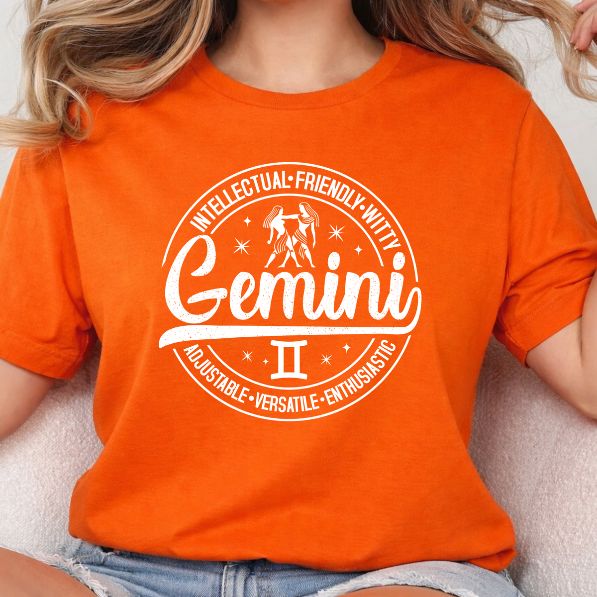 orange t-shirt laid flat, featuring a circular white Gemini zodiac graphic with the twins illustration, the word "Gemini," and trait keywords like intellectual, friendly, witty, enthusiastic, versatile, and adjustable.