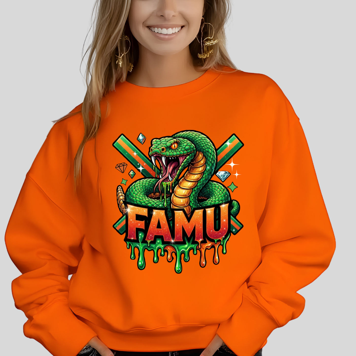 A woman wearing a bright orange crewneck sweatshirt with a colorful graphic of a green snake, crossed green bats, diamonds, and the text 'FAMU' in dripping orange and green letters.