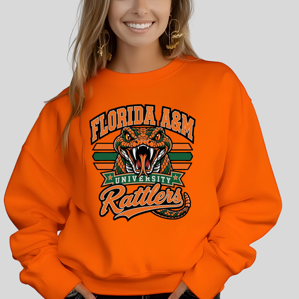 A woman wearing a bright orange crewneck sweatshirt with a colorful graphic of a green snake, crossed green bats, diamonds, and the text 'FAMU' in dripping orange and green letters.