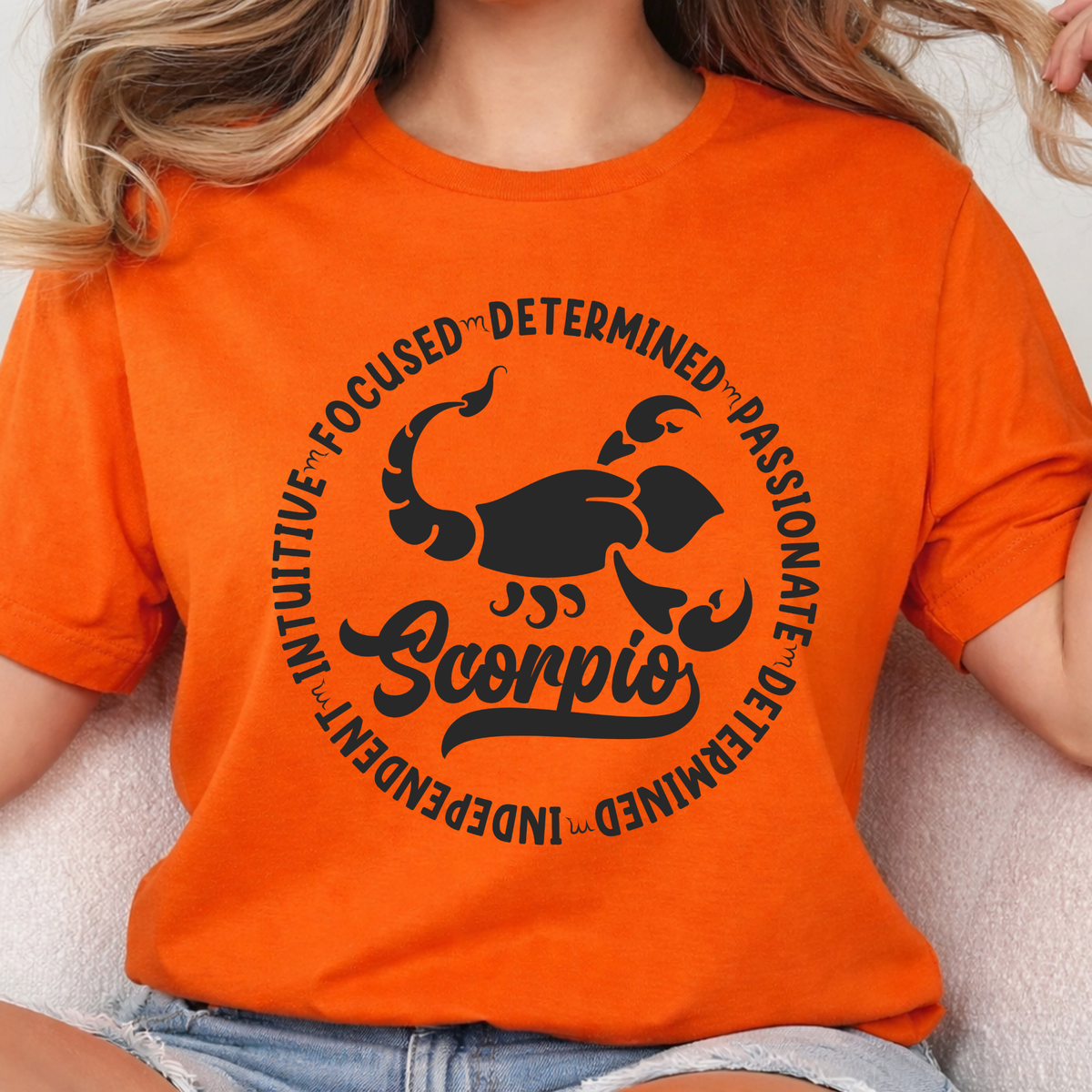 Woman wearing a orange crewneck sweatshirt featuring a white circular Scorpio zodiac graphic with a scorpion illustration, the word "Scorpio," and trait keywords like intuitive, focused, determined, passionate, and independent.