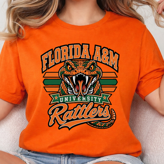 A person wearing a bright orange graphic T-shirt featuring a green coiled snake, crossed green swords, diamonds, and the word "FAMU" in dripping orange and green lettering.