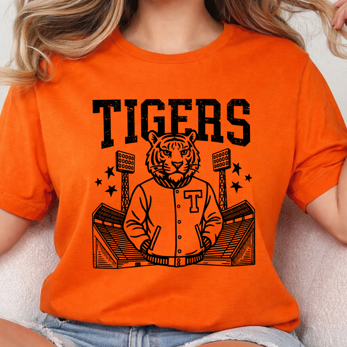 A woman wearing a bright orange short-sleeve T-shirt with a black graphic of a tiger mascot in a varsity jacket playing drums in front of a stadium, with the word 'TIGERS' above. The photo is cropped from the shoulders to the hips against a light neutral background.