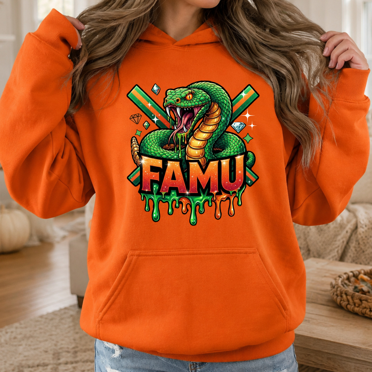 A woman wearing a bright orange hoodie with a colorful graphic of a green snake, crossed green and orange sticks, diamonds, and the word 'FAMU' in dripping letters.