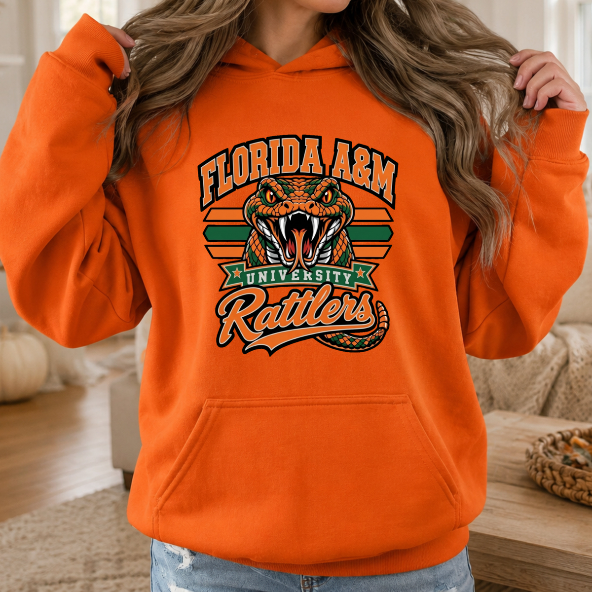 A woman wearing a bright orange hoodie with a colorful graphic of a green snake, crossed green and orange sticks, diamonds, and the word 'FAMU' in dripping letters.