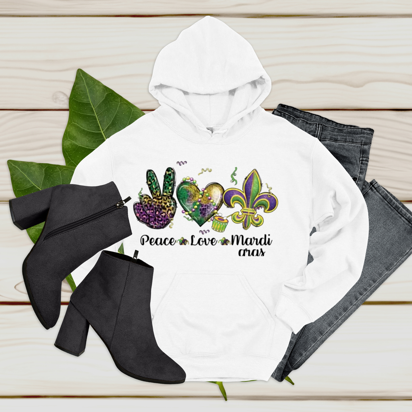 White hoodie with Mardi Gras-themed design, black boots, and jeans on a wooden surface.
