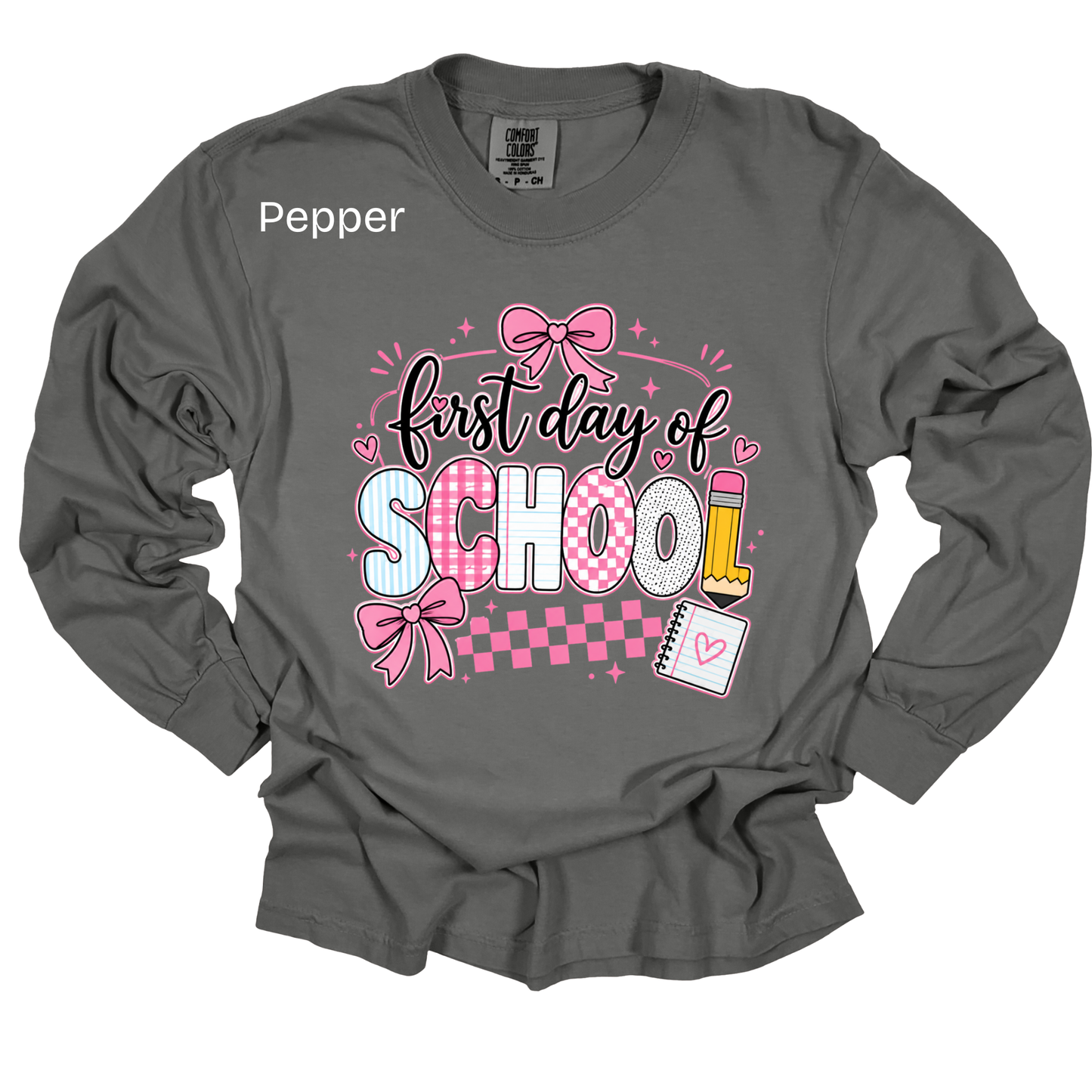 A dark gray long-sleeve t-shirt with a 'first day of SCHOOL' graphic on the front. The graphic features pink and white checkered lettering, pink bows, a yellow pencil, and a small notebook.