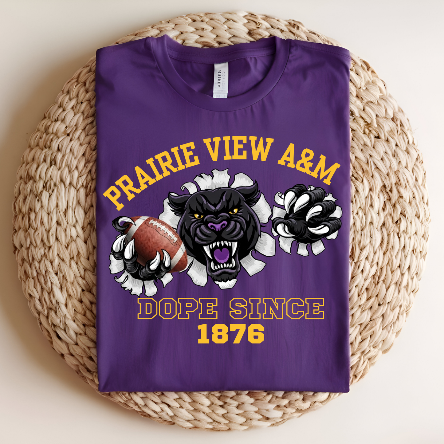 A folded purple graphic T-shirt displayed on a round woven straw mat. The shirt features a bold design with the text 'PRAIRIE VIEW A&M' and 'DOPE SINCE 1876' in yellow, along with a black panther mascot holding a football and white flower-like shapes.