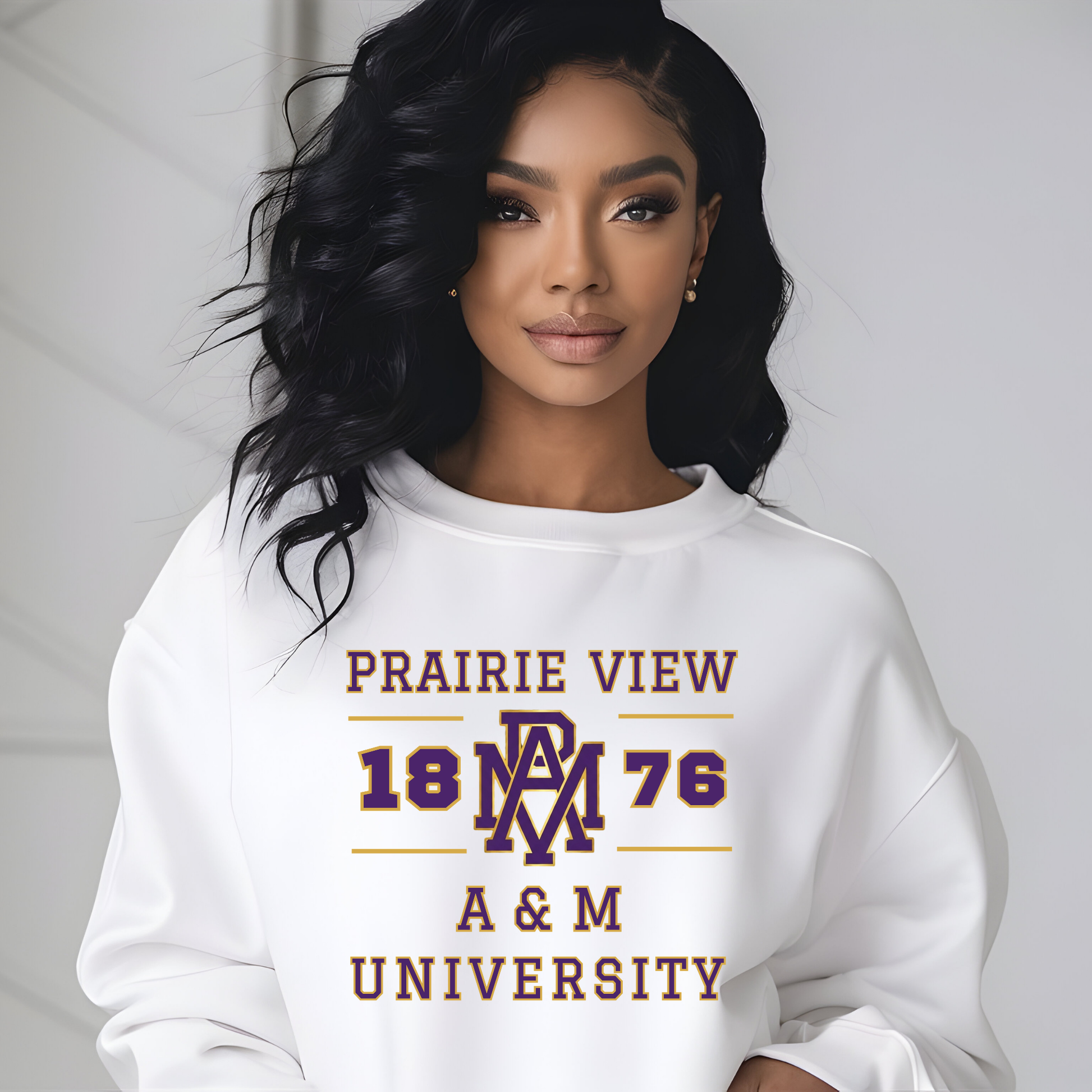 A woman with long, wavy black hair is wearing a white crewneck sweatshirt featuring a maroon and gold graphic that reads 'PRAIRIE VIEW 1876 A & M UNIVERSITY' with a stylized 'PW' logo. The background is a clean, light gray wall.
