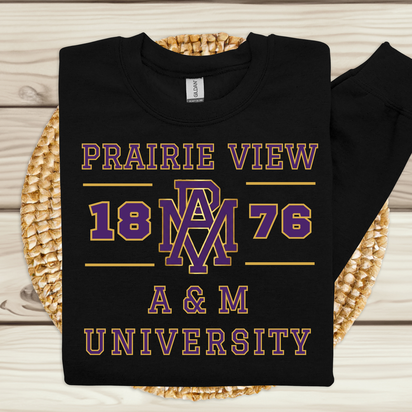 A black long-sleeve t-shirt with a purple and gold graphic print reading 'PRAIRIE VIEW 1876 A & M UNIVERSITY' and an 'AM' monogram, laid flat on a woven round mat over a light wood plank background.