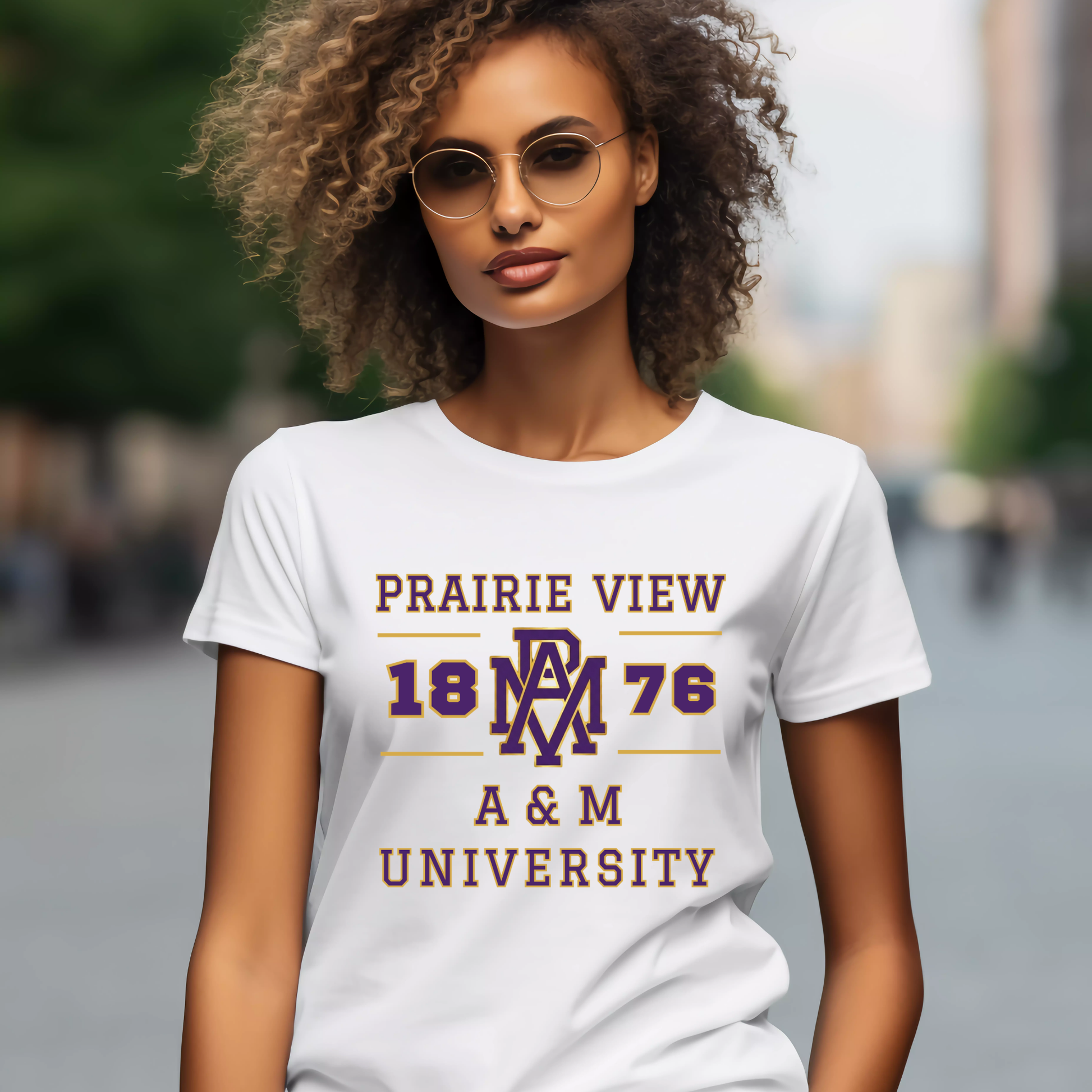 A woman wearing round sunglasses and a white graphic T-shirt with 'PRAIRIE VIEW 1876 A&M UNIVERSITY' printed in maroon and purple on the front, photographed outdoors with a blurred city background.