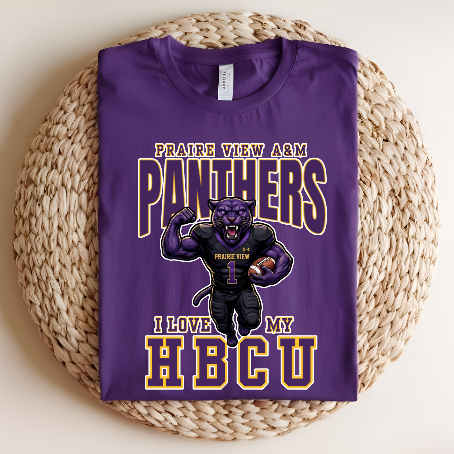 A folded purple graphic T-shirt displayed on a round woven straw mat. The shirt features a cartoon panther mascot in a black football uniform with the text 'PRAIRIE VIEW A&M PANTHERS' and 'I LOVE MY HBCU' in large outlined lettering.