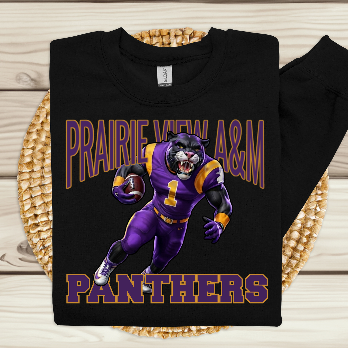 A black long-sleeve sweatshirt laid flat on a woven round mat over a light wood surface. The sweatshirt features a large graphic of a purple and gold football mascot running, with the text 'PRAIRIE VIEW A&M' above and 'PANTHERS' below in purple and gold.