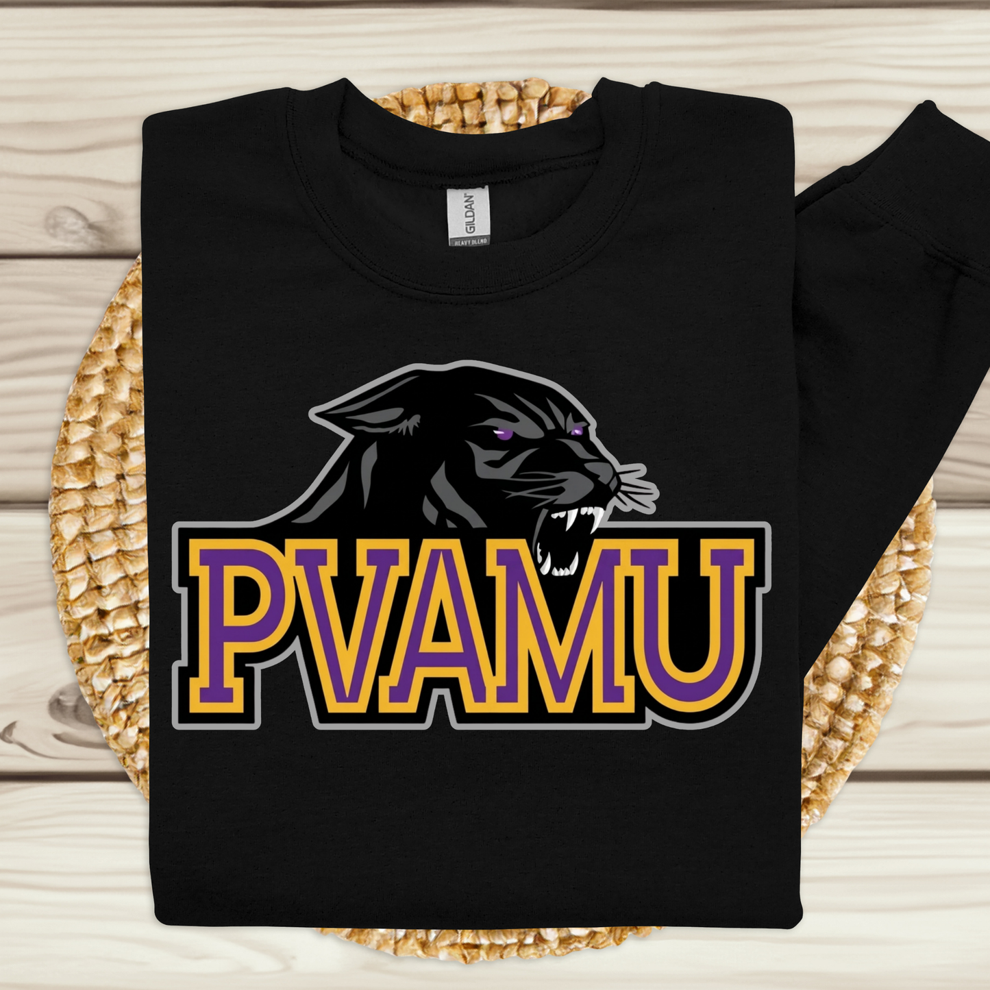 A black long-sleeve t-shirt with a graphic of a black panther and the text 'PVAMU' in purple and gold, displayed on a woven placemat over a light wood background.
