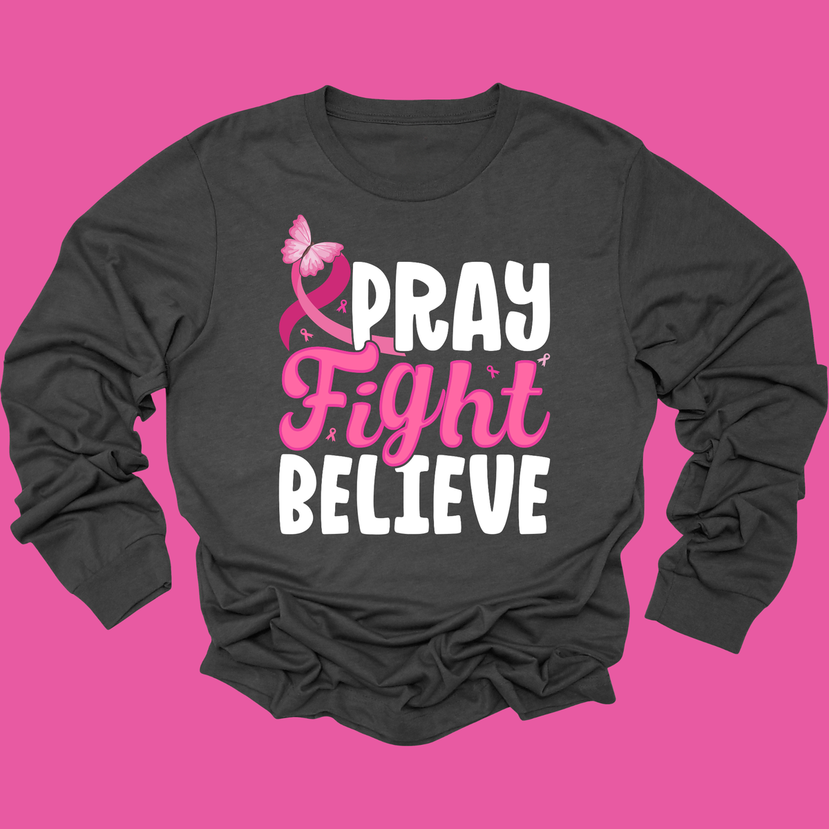 A dark charcoal gray long-sleeve t-shirt with a pink and white graphic reading 'PRAY Fight BELIEVE' with a pink ribbon and butterfly, displayed on a solid bright pink background.