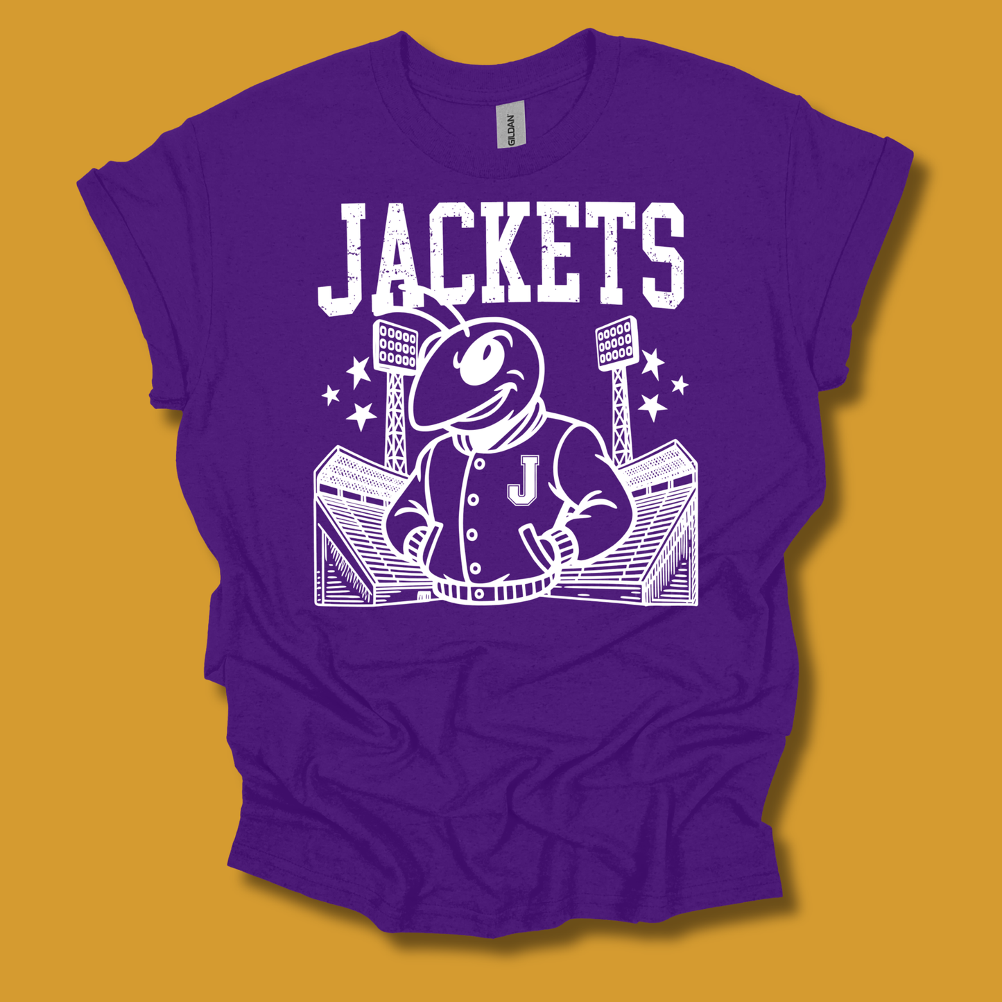 A purple short-sleeve graphic T-shirt laid flat against a mustard yellow background. The shirt features a large white printed design with the word "JACKETS" above a cartoon mascot in a jacket standing in front of a stadium, with a small "Gildan" neck label visible inside the collar.