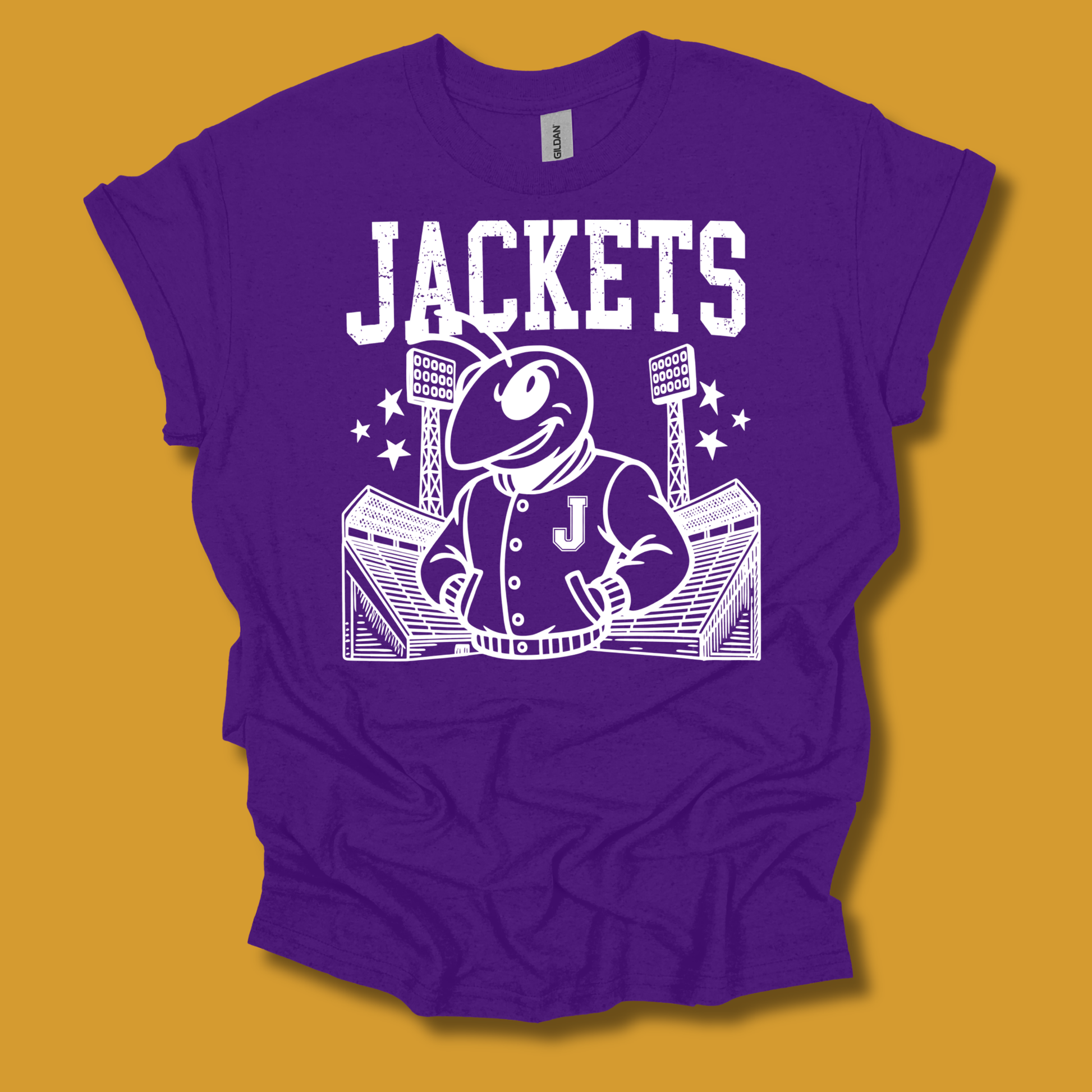 A purple short-sleeve graphic T-shirt laid flat against a mustard yellow background. The shirt features a large white printed design with the word "JACKETS" above a cartoon mascot in a jacket standing in front of a stadium, with a small "Gildan" neck label visible inside the collar.