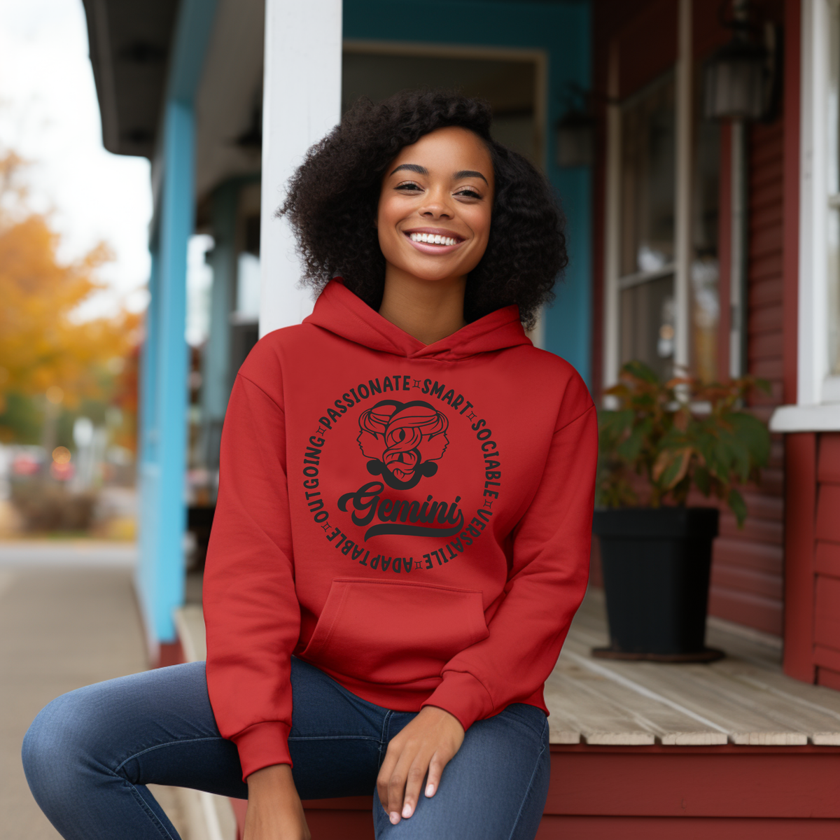 A smiling woman with curly dark hair sits on a porch step wearing a bright red pullover hoodie with a black circular Gemini graphic print on the front, paired with blue jeans.