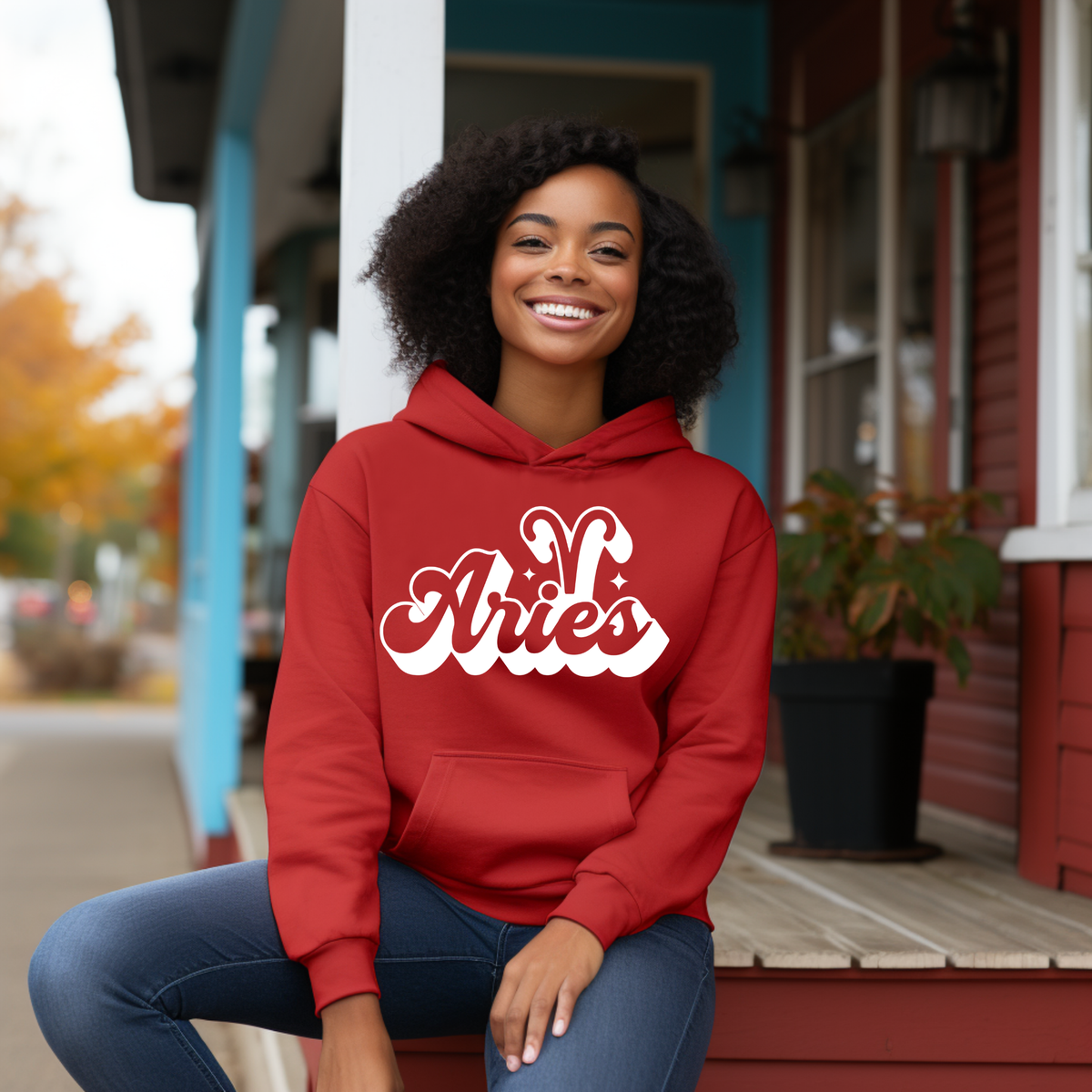 A woman with curly black hair sits on the wooden porch steps of a house, wearing a red hoodie with 'Aries' written in white cursive script across the chest, paired with blue jeans.