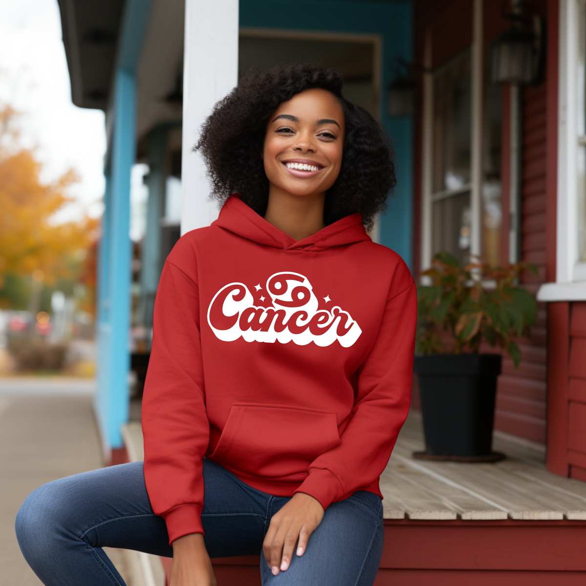 A smiling woman with curly dark hair sits on a porch step wearing a red hoodie with 'Cancer' printed in white script across the chest. The background shows a residential house porch with blue trim, a potted plant, and blurred outdoor scenery.