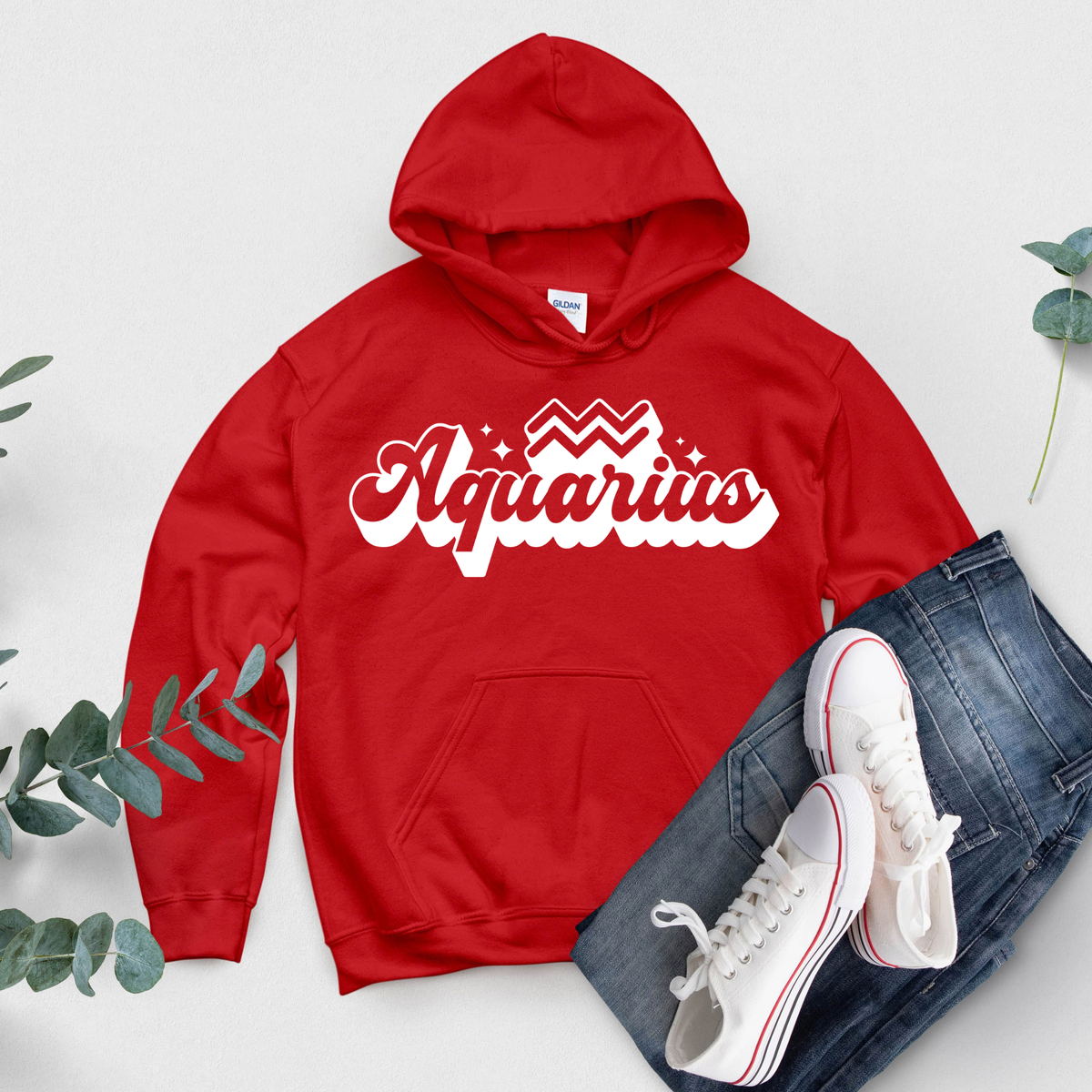 A red pullover hoodie with 'Aquarius' printed in white script across the chest, styled in a flat lay with blue jeans, white sneakers, and green eucalyptus leaves on a light gray background.