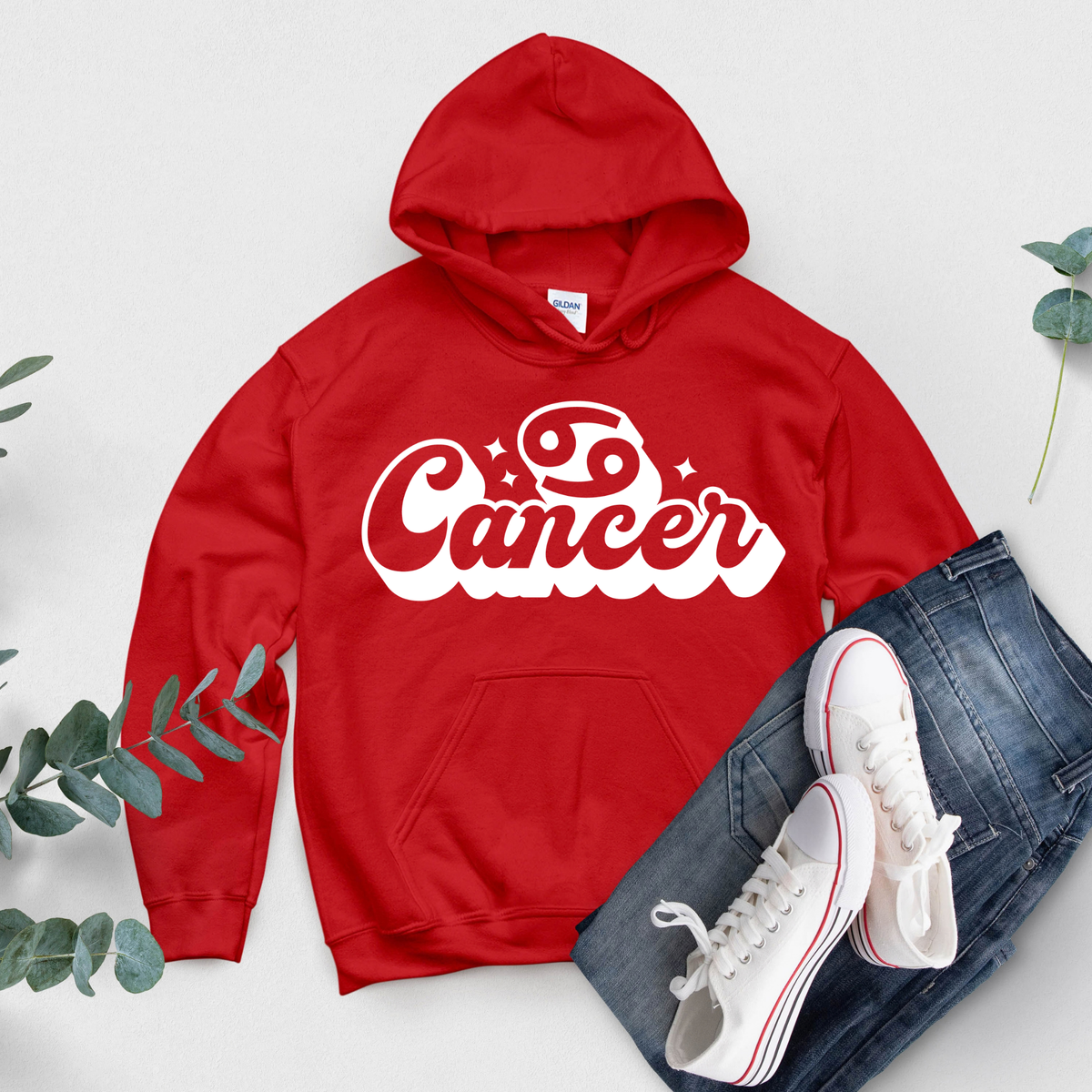 A red pullover hoodie with a white 'Cancer' zodiac graphic on the chest, styled in a flat lay with blue jeans, white sneakers, and green eucalyptus leaves on a light gray background.