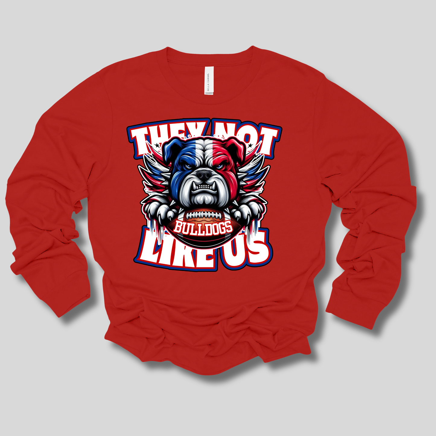 A red long-sleeve t-shirt with a graphic of a bulldog mascot holding a football, with the text 'THEY NOT LIKE US' and 'BULLDOGS' in red, white, and blue.