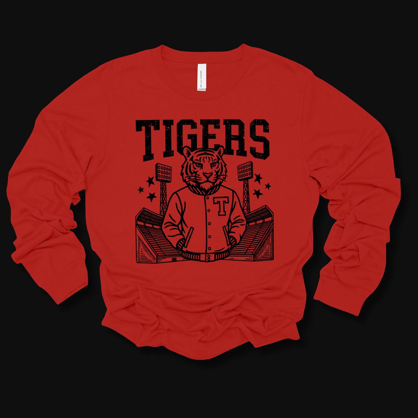 A red long-sleeve t-shirt with a black graphic print of a tiger mascot wearing a varsity jacket in a stadium, with the word 'TIGERS' printed above. The shirt is laid flat on a solid black background.