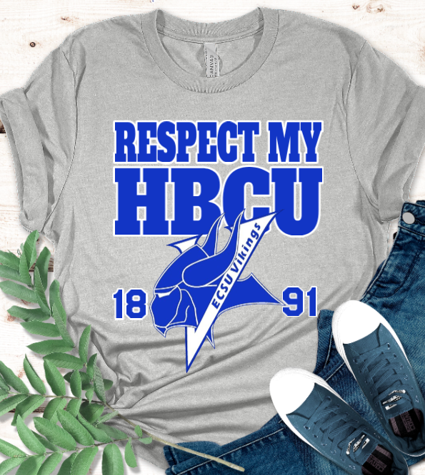A heather gray graphic T-shirt laid flat on a light wood surface, styled with blue jeans, navy sneakers, green leaves, and a small plant sprig. The shirt has large blue text reading 'RESPECT MY HBCU' with a blue mascot graphic and '1891' and 'Vikings' details.