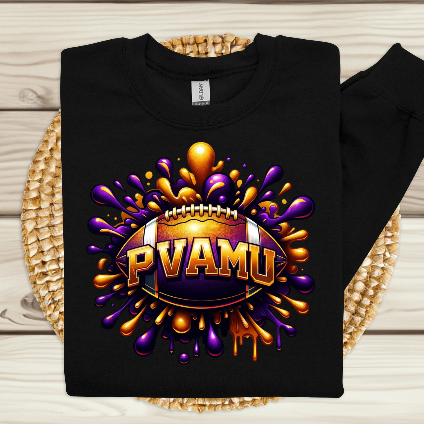 A black long-sleeve sweatshirt laid flat on a woven placemat over a light wood surface. The front has a large colorful graphic of a football surrounded by purple and gold paint splatters, with the text 'PVAMU' across the center.