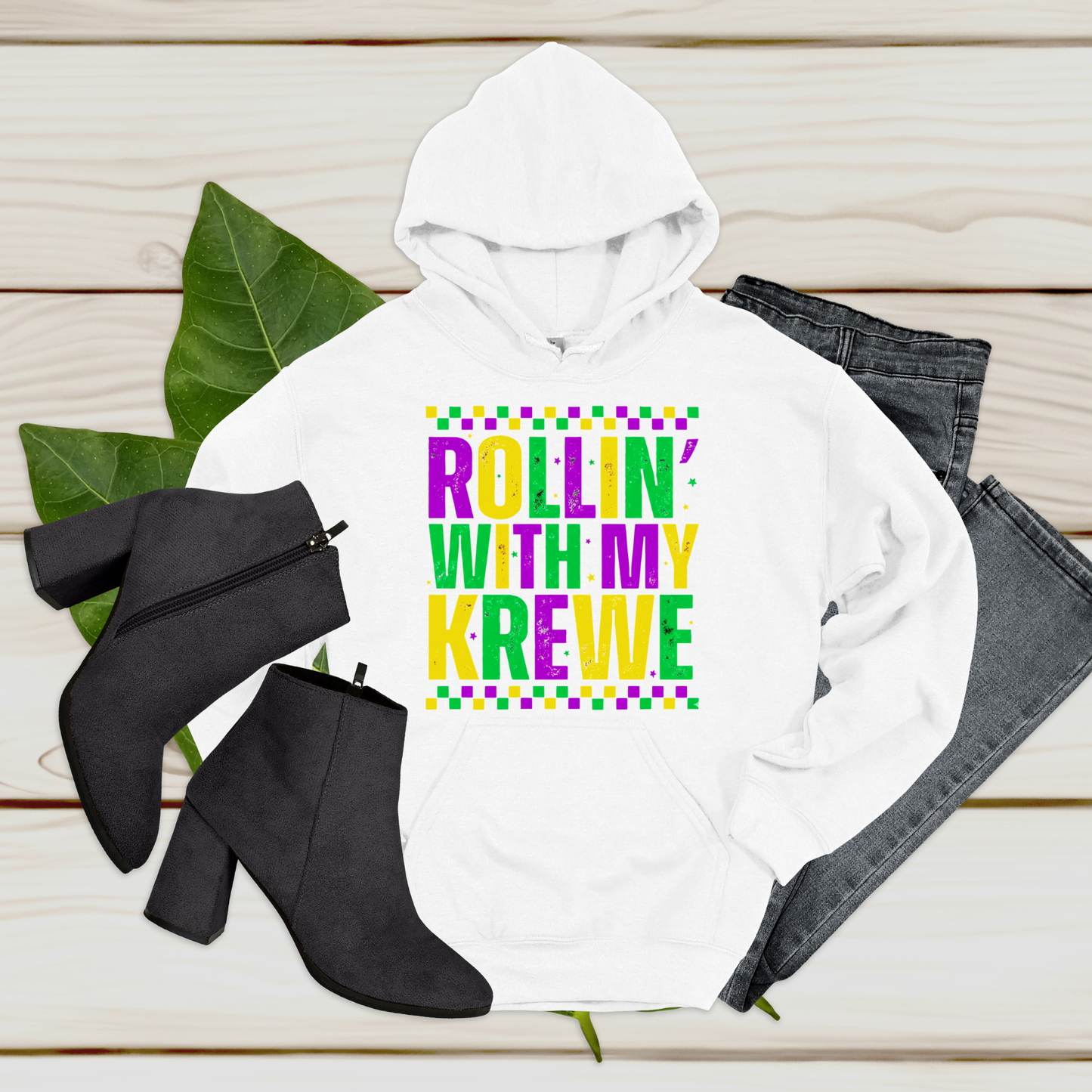 White hoodie with colorful text 'Rollin' with my Krewe', black boots, and jeans on a wooden surface.