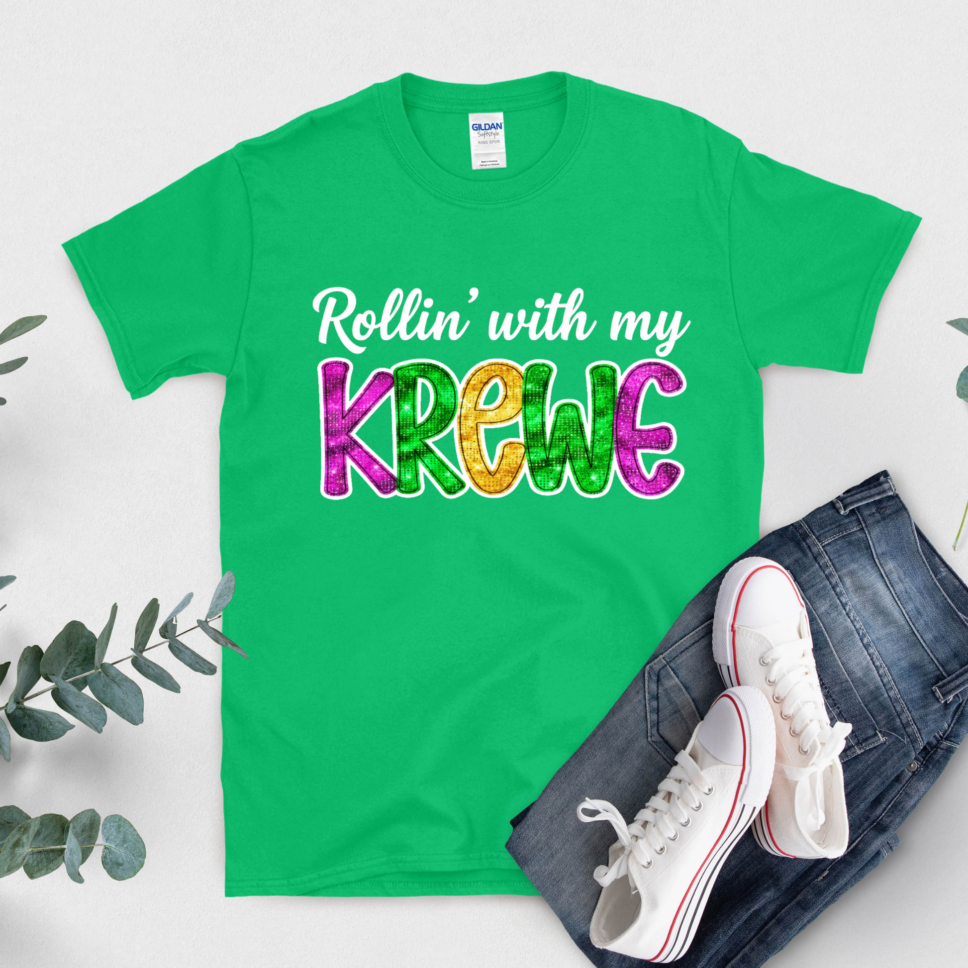 Green t-shirt with colorful text 'Rollin' with my Krewe', white sneakers, and jeans on a light gray background.