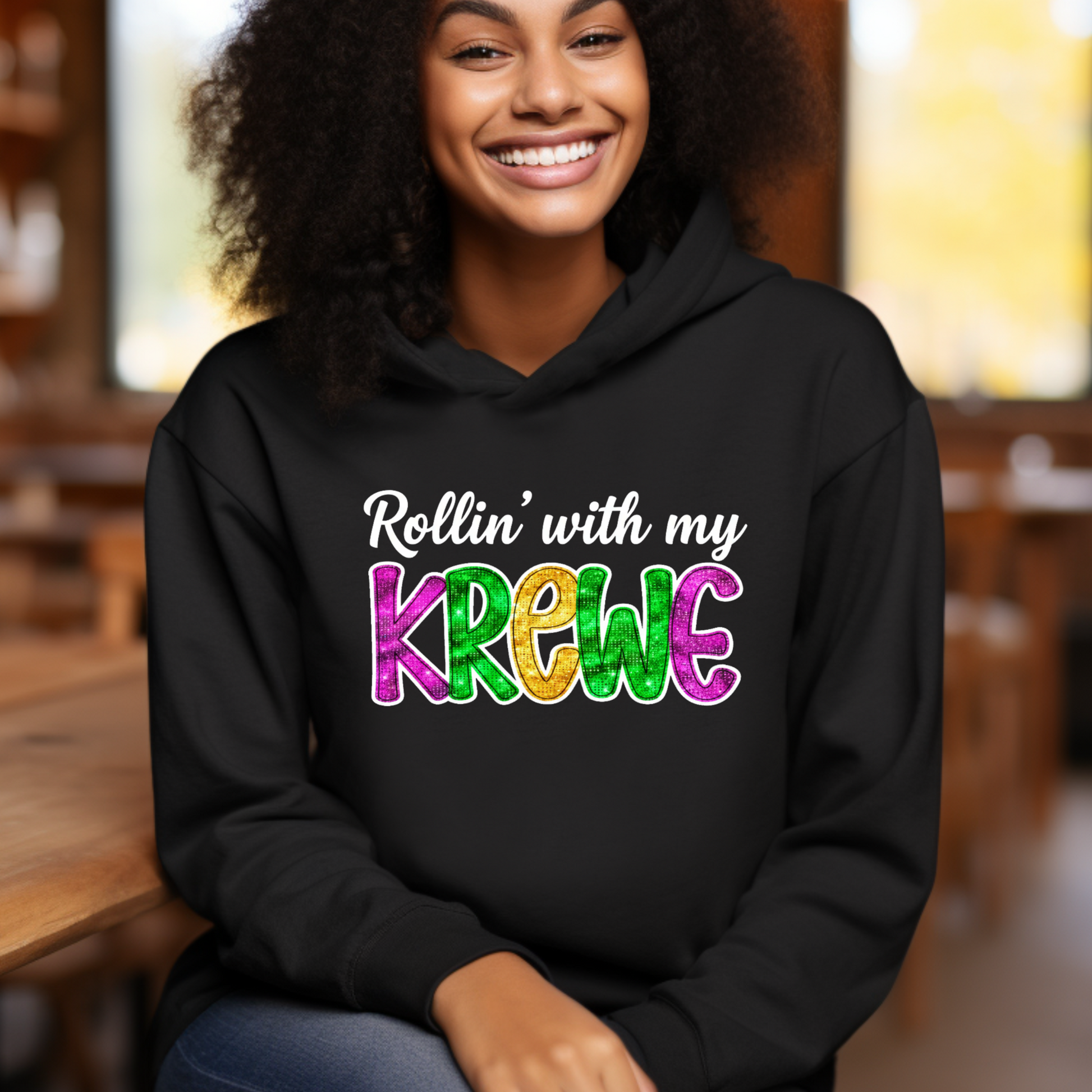 Person wearing a black hoodie with 'Rollin' with my Krewe' text in colorful letters, sitting indoors.