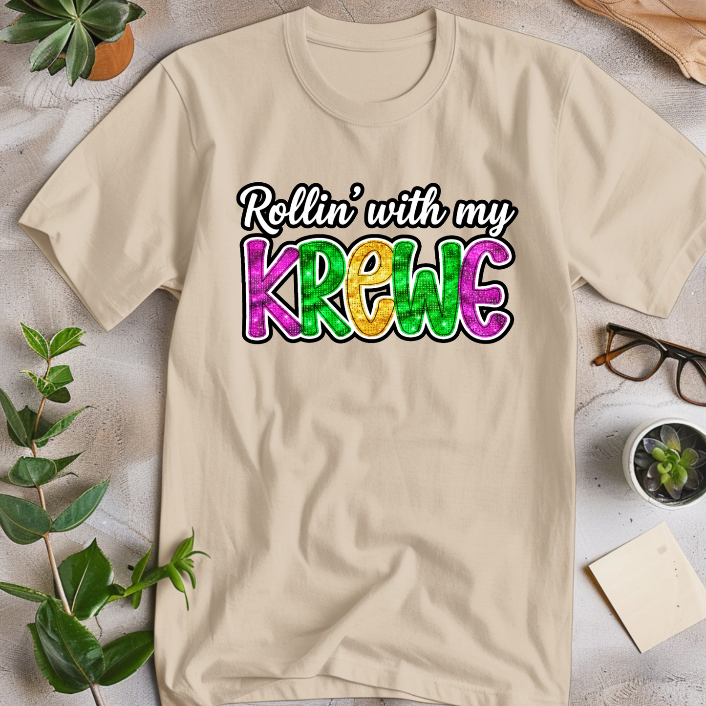 Beige t-shirt with 'Rollin' with my Krewe' text on a light surface with plants and glasses.