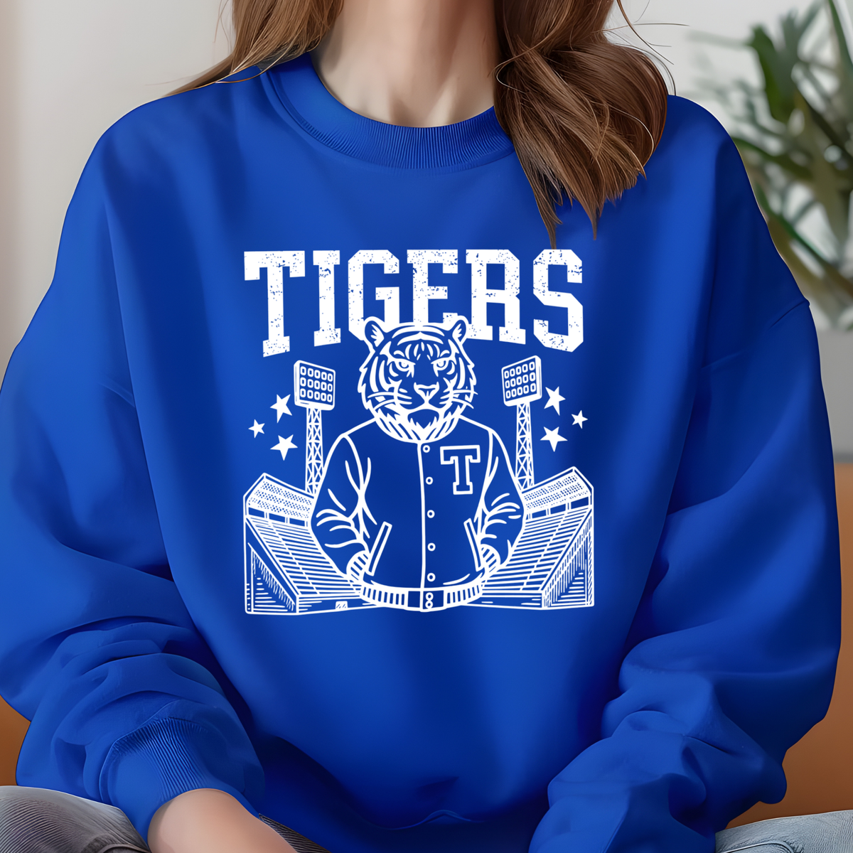 A person wearing a royal blue crewneck sweatshirt with a white graphic of a tiger mascot in a baseball jacket and stadium, with the word 'TIGERS' above.