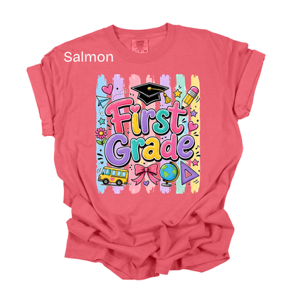 A salmon pink short-sleeve t-shirt with a colorful 'First Grade' graphic on the front, featuring school-themed doodles like a graduation cap, pencil, globe, and bus. The word 'Salmon' is printed in white text near the upper left chest.