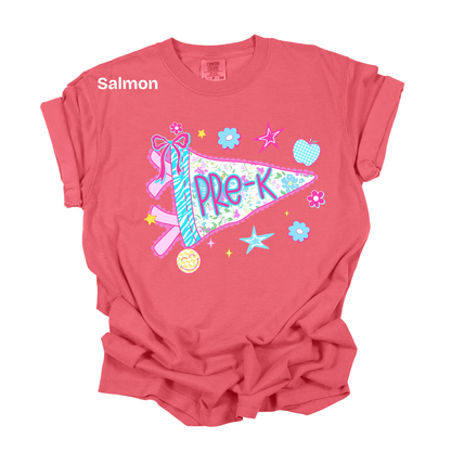 A salmon pink short-sleeve t-shirt with a colorful graphic on the front featuring a ribbon, flowers, stars, and a pennant that reads 'PRE-K'. The word 'Salmon' is printed in white on the upper left chest.