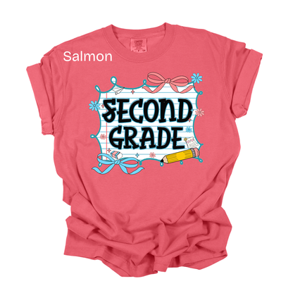 A salmon pink short-sleeve t-shirt with a 'SECOND GRADE' school-themed graphic on the front, featuring a blue and white grid frame, pink bows, and a yellow pencil. The word 'Salmon' is printed in white text near the upper left chest.