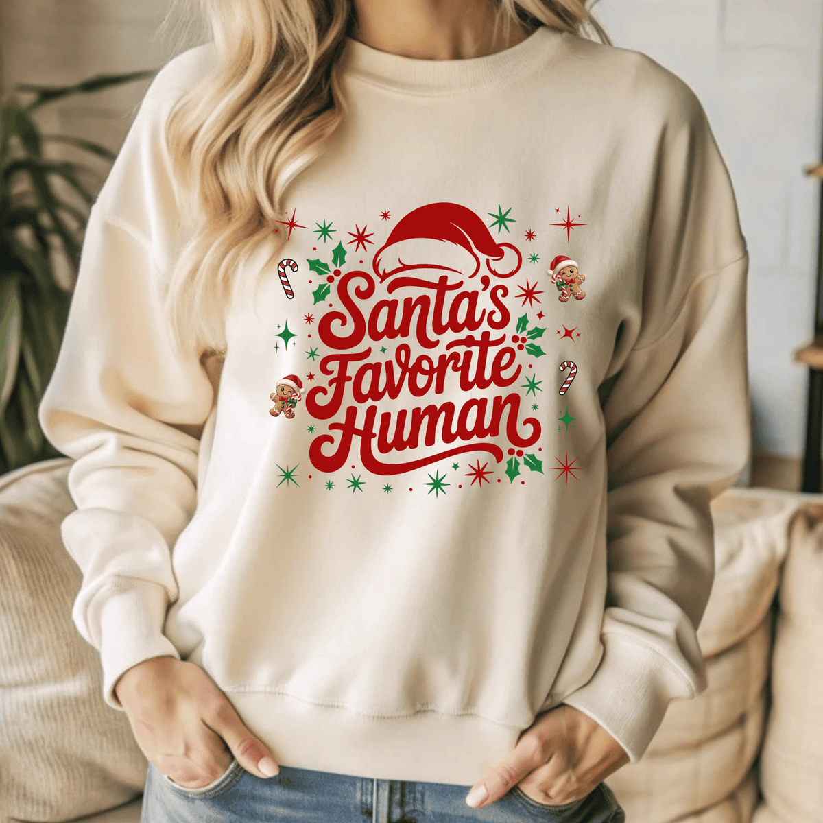 Folded sand crewneck sweatshirt with 'Santa's Favorite Human' red lettering, Santa hat, and gingerbread graphics on a neutral wooden background.