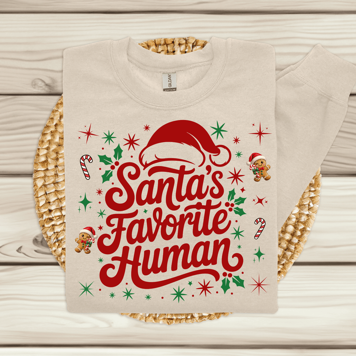 Folded sand crewneck sweatshirt with 'Santa's Favorite Human' red lettering, Santa hat, and gingerbread graphics on a neutral wooden background.