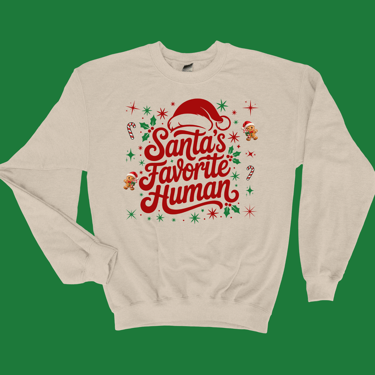 Folded sand crewneck sweatshirt with 'Santa's Favorite Human' red lettering, Santa hat, and gingerbread graphics on a neutral wooden background.
