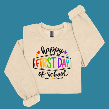 A cream-colored long-sleeve sweatshirt with a colorful 'happy first day of school' graphic on the front, displayed against a solid teal-blue background.