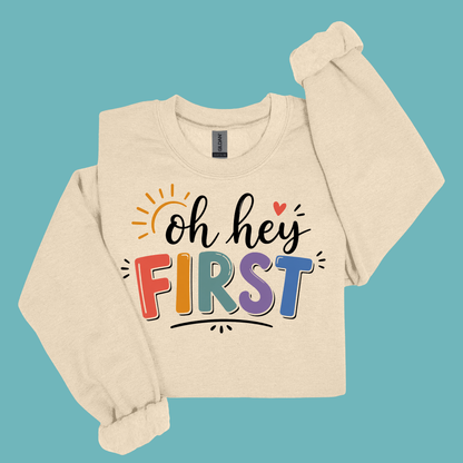 A beige long-sleeve sweatshirt with a colorful graphic reading 'Kinder Garten' in decorative letters, surrounded by small Easter-themed illustrations and a pink bow, shown on a solid teal background.