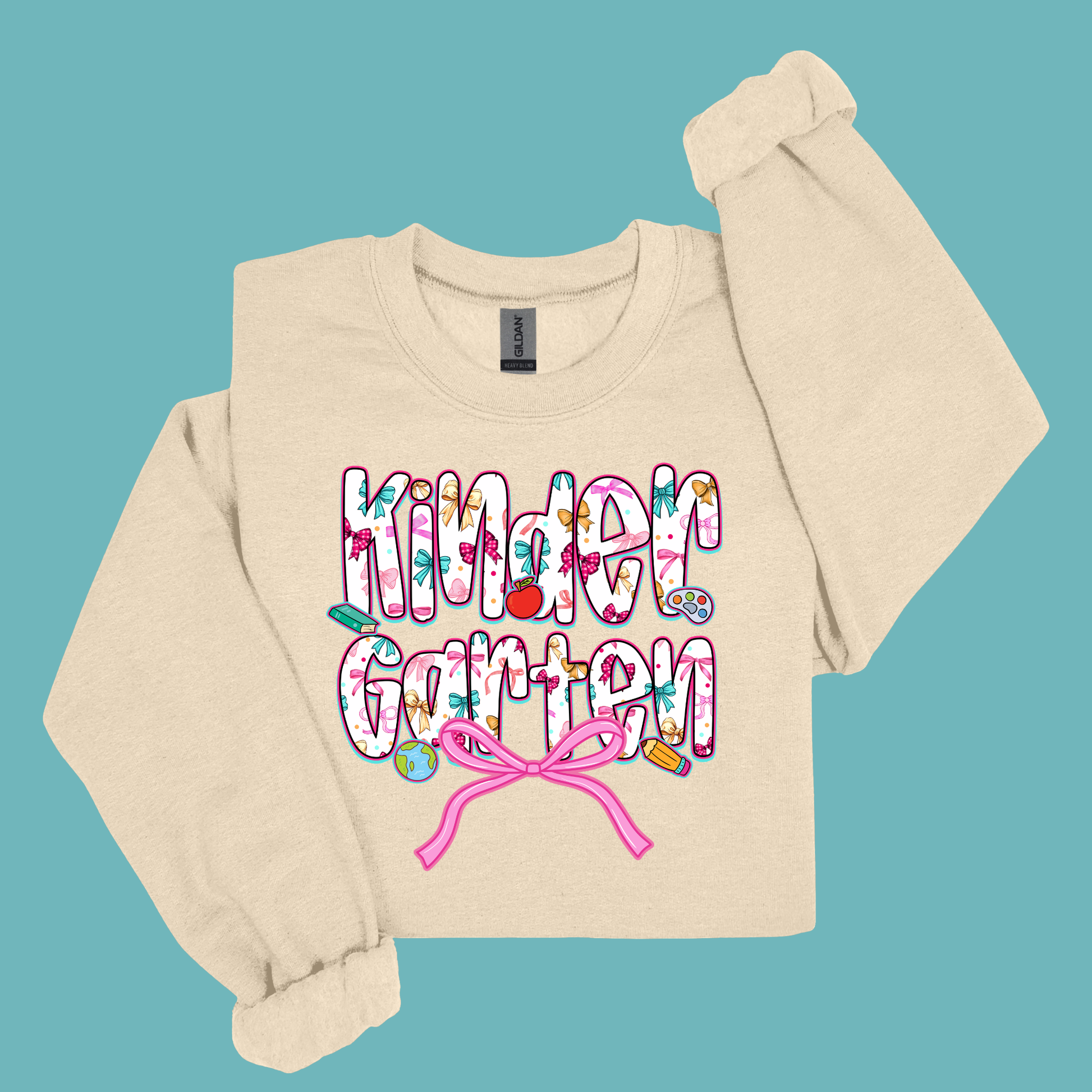 A beige long-sleeve sweatshirt with 'Kinder Garten' printed in a colorful, playful font on the front, featuring a pink bow and Easter-themed doodles, displayed on a solid teal background.