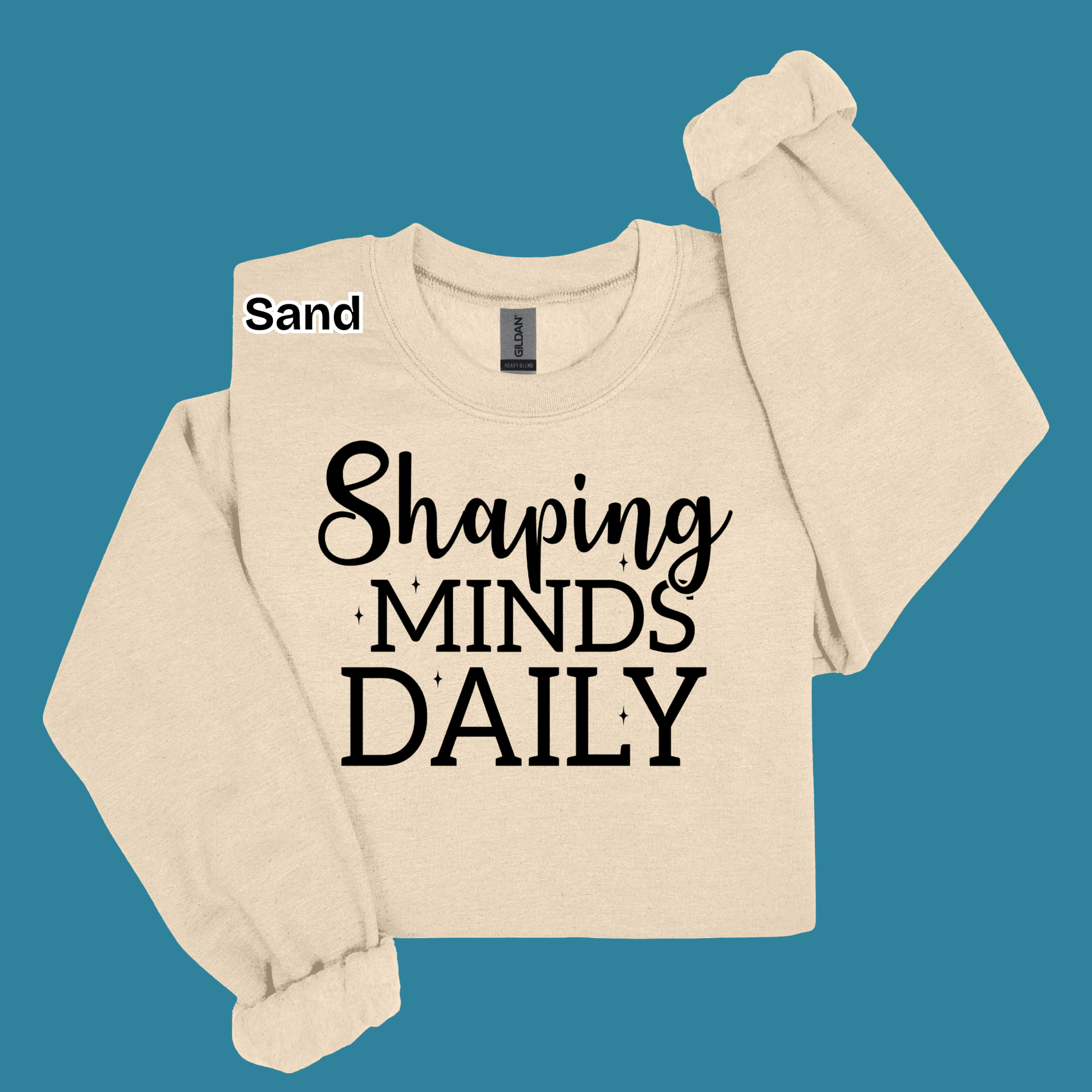 A cream-colored long-sleeve sweatshirt with the text 'Shaping MINDS DAILY' printed in black on the front, displayed against a solid teal background. A small label reading 'Sand' is visible near the collar.