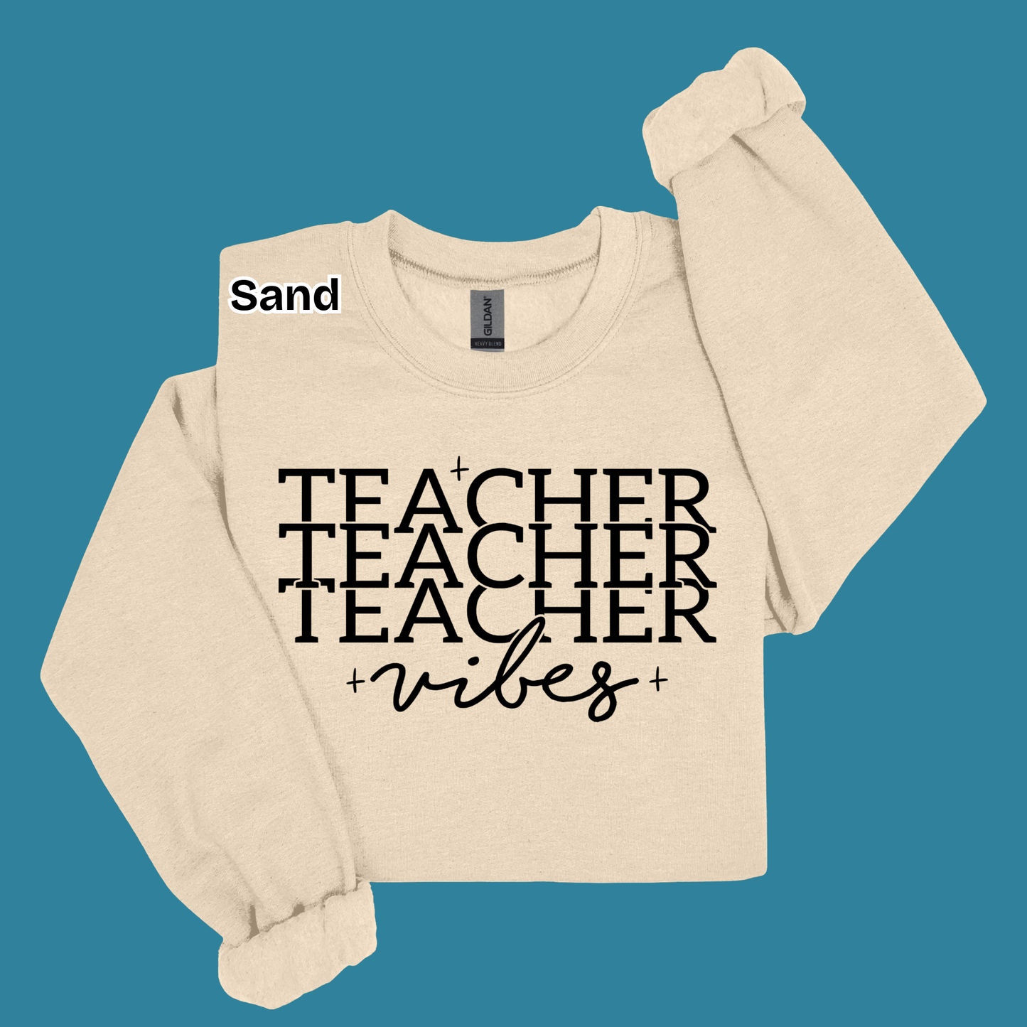 A beige crewneck sweatshirt with 'TEACHER TEACHER TEACHER' printed in black block letters and 'vibes' in black script below it, shown on a solid teal background with a 'Sand' color label.