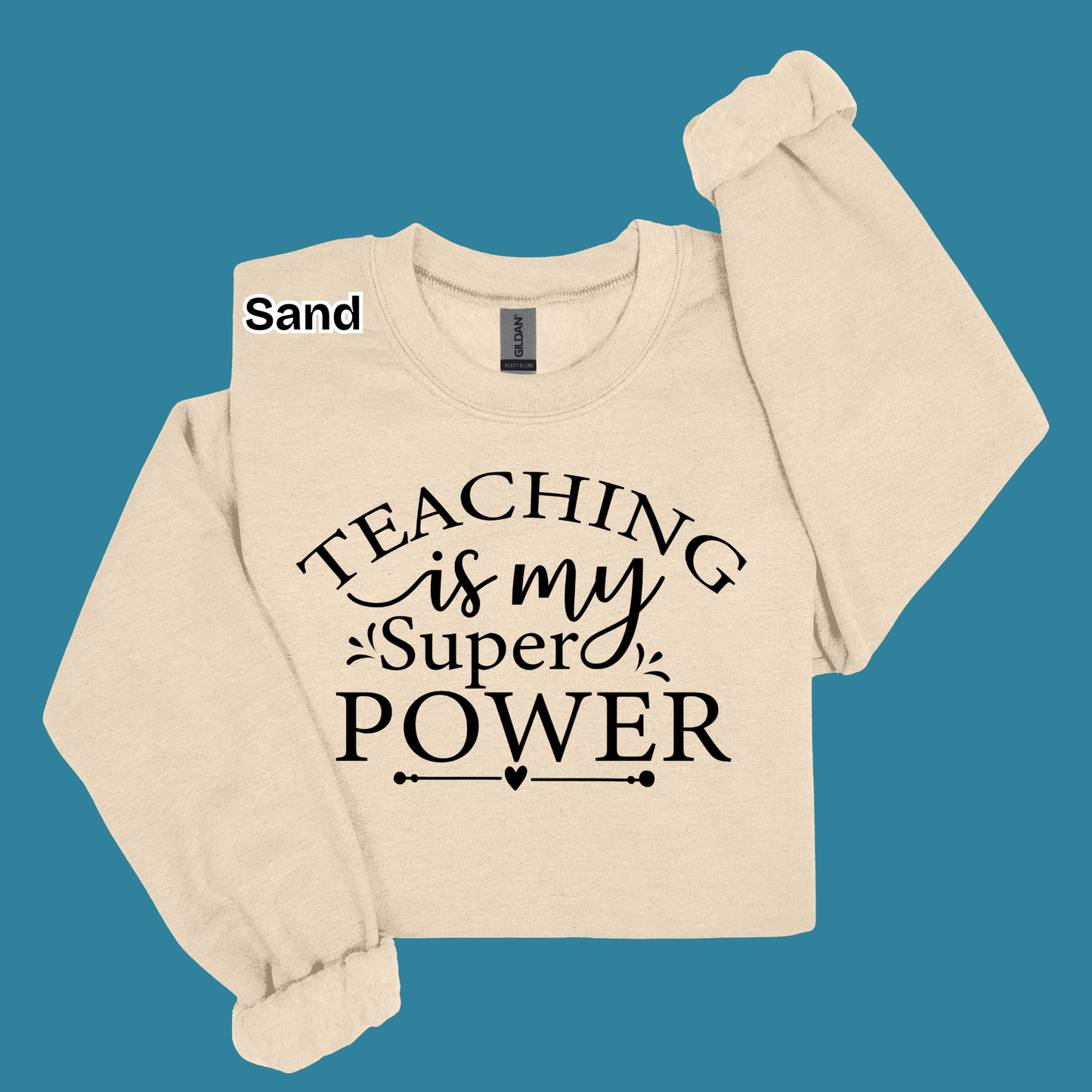 A cream-colored long-sleeve sweatshirt with 'TEACHING is my Super POWER' printed in black on the front, displayed on a solid teal background. A small label near the collar reads 'Sand'.